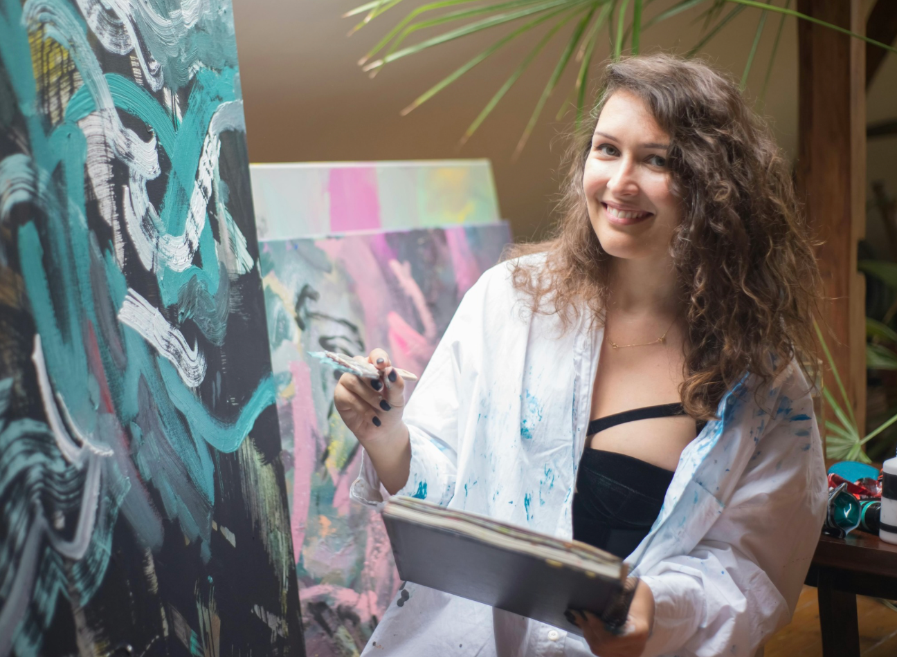 Smiling woman with white man's shirt on sitting in front of an easel brush in hand ready to paint.